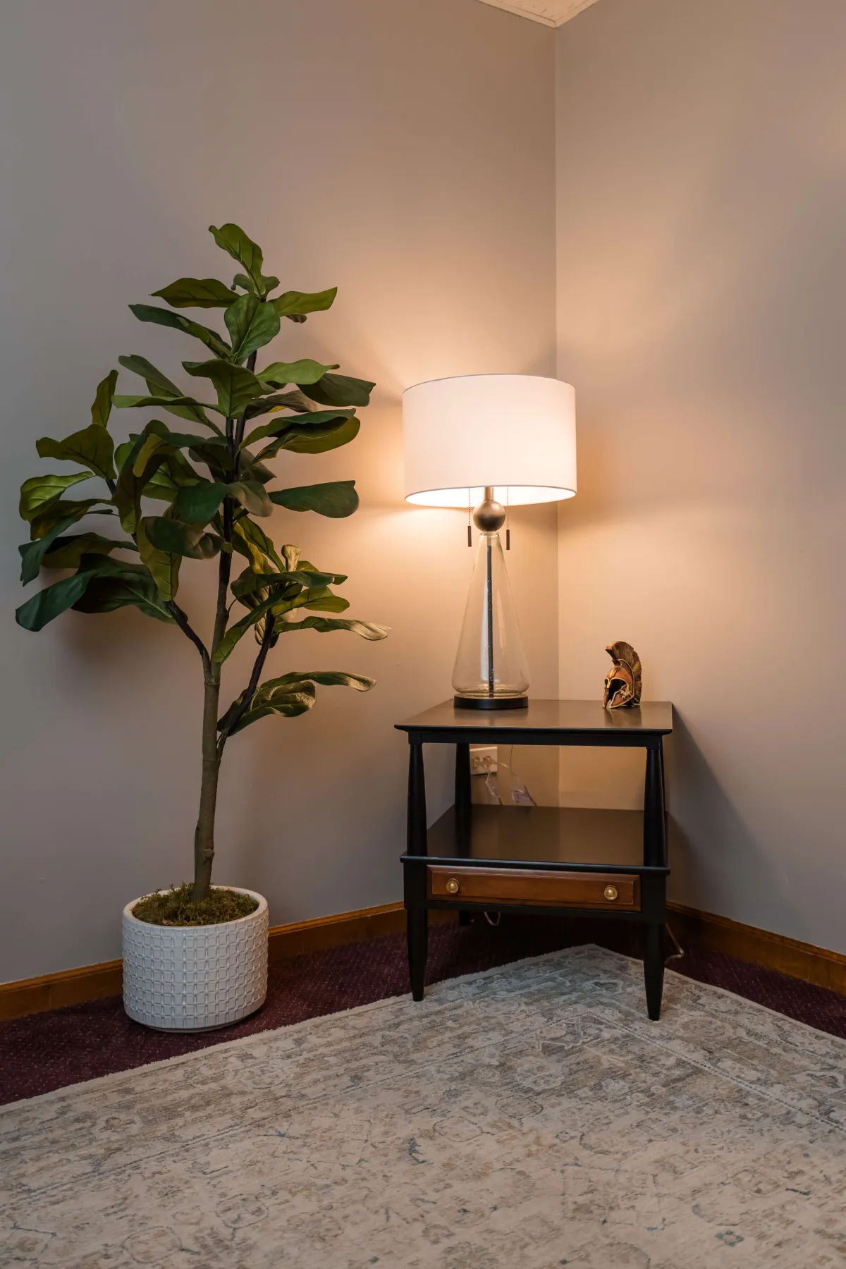 Lamp sits on table in room corner next to large fiddle leaf fig tree.